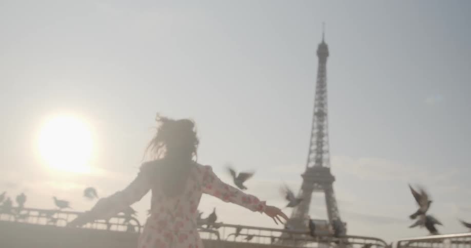 A woman in a floral dress playfully runs towards a flock of pigeons on a wall in view of the eiffel tower