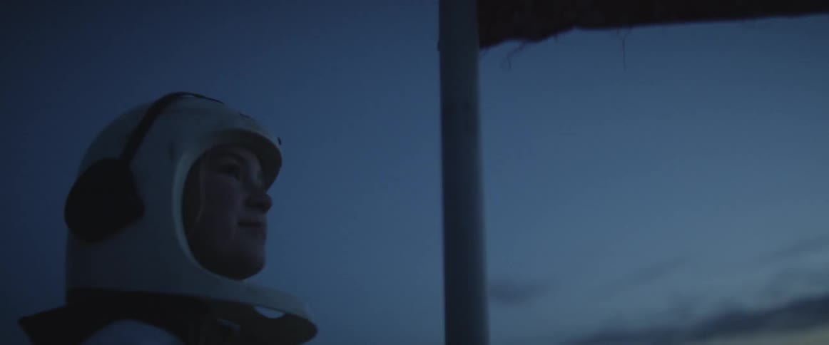 A young female wears a costume astronaut helmet and outfit as she drive a flag pole into the ground an looks out over the distance