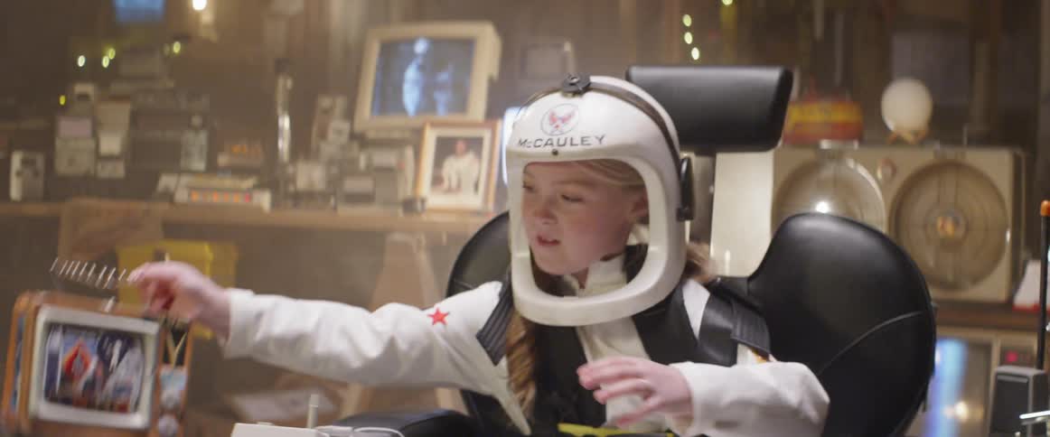 A young girl in an astronaut costume plays with a fake control board and grabs a lunch box