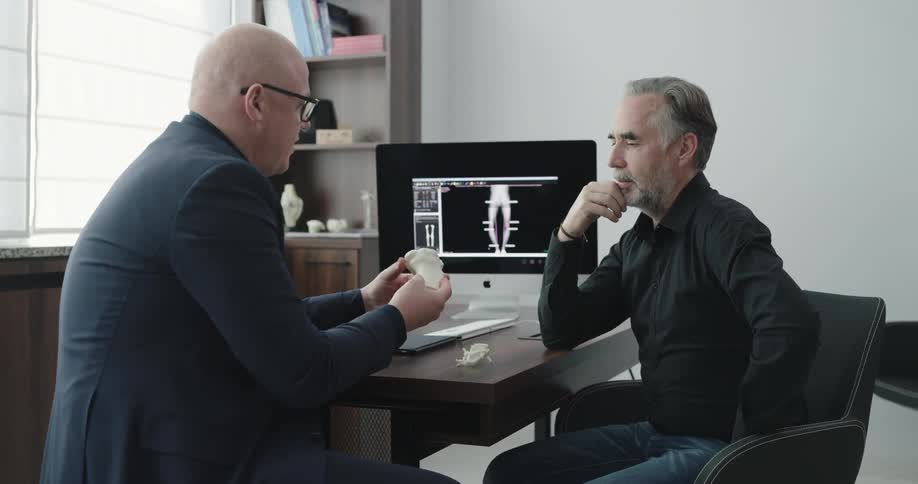 Two men sit at a desk talking while one man shows him how bones go together