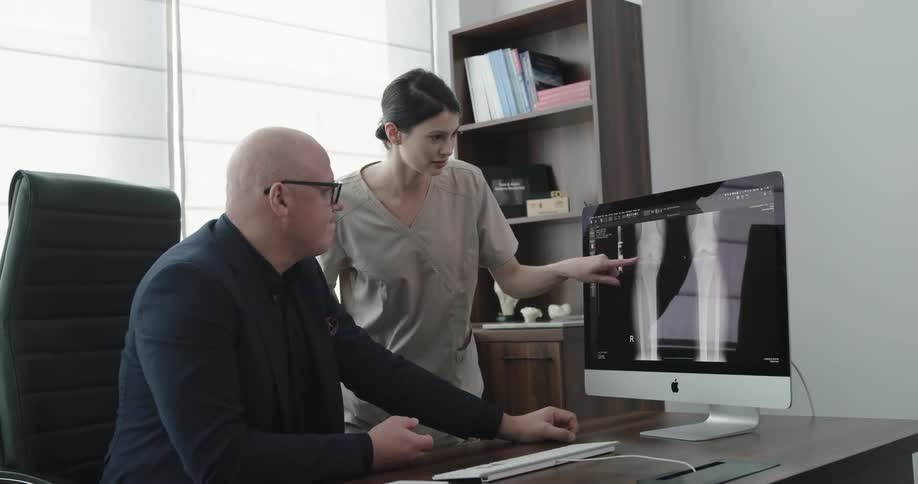 A man sits at a desk discussing an image of a joint on his screen with the woman standing next to him