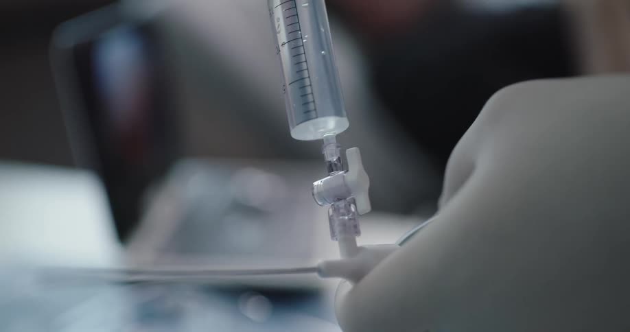 A person wearing a latex glove holds a needle as they push a clear liquid through it before turning towards a patient