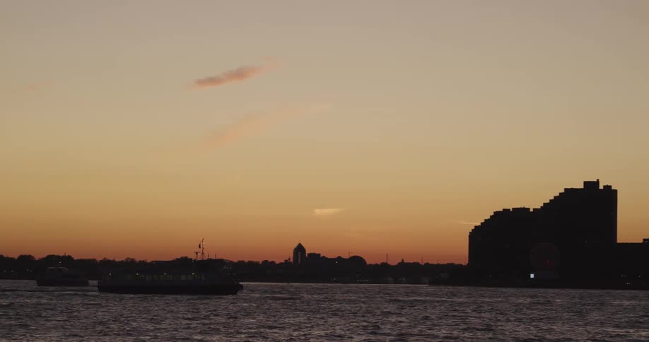 A boat sails down a river until it passes by a large building by the waterfront at dusk