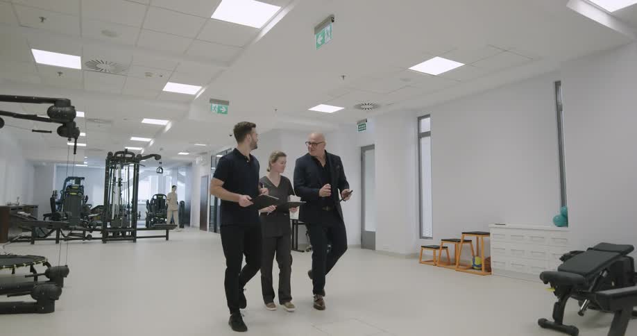 While discussing their notes three people walk through a physical therapy facility that has a variety of exercise equipment