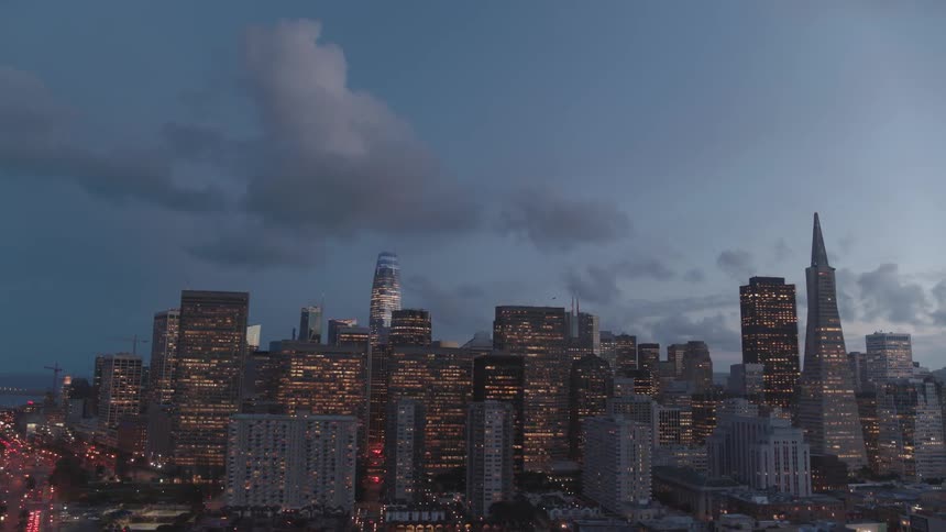 Clouds hang in the sky above a city skyline with lights glowing in skyscraper windows and on roadways