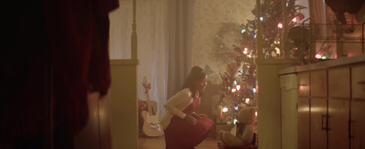 A black woman with straightened hair kneels by a christmas tree and then walks away