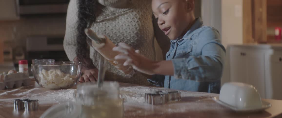 A young black girl is using a rolling pin to roll dough on a floured counter while an adult stands beside her watching