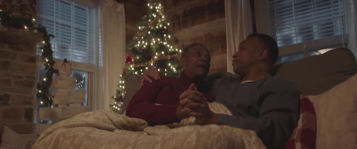 Male and female sit together on a love seat covered with a blanket while talking and holding hands in a room decorated for christmas