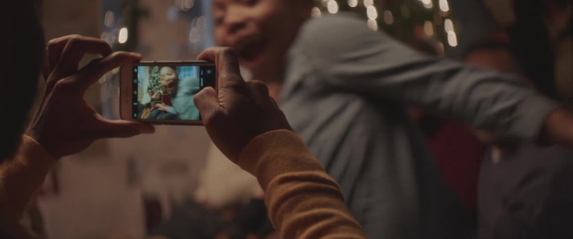 Black man takes a picture of the black family sitting on the couch next to the christmas tree with a smartphone at home