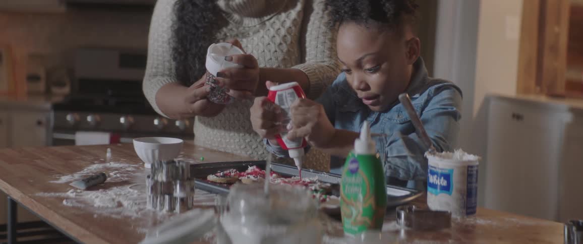 A mother and daughter decorate sugar cookies with icing and sprinkles in a home kitchen