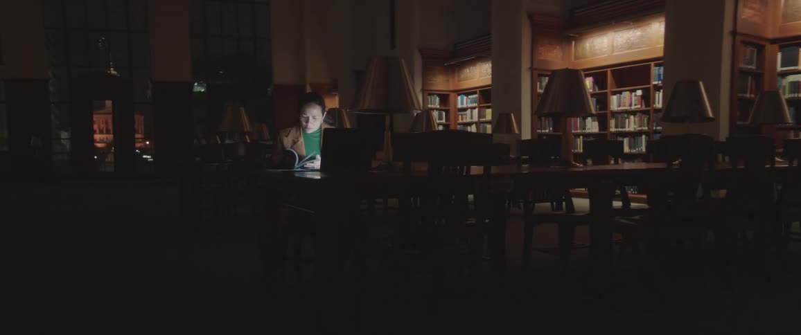 Woman sitting at the table studies and use laptop while people walk in the library at night