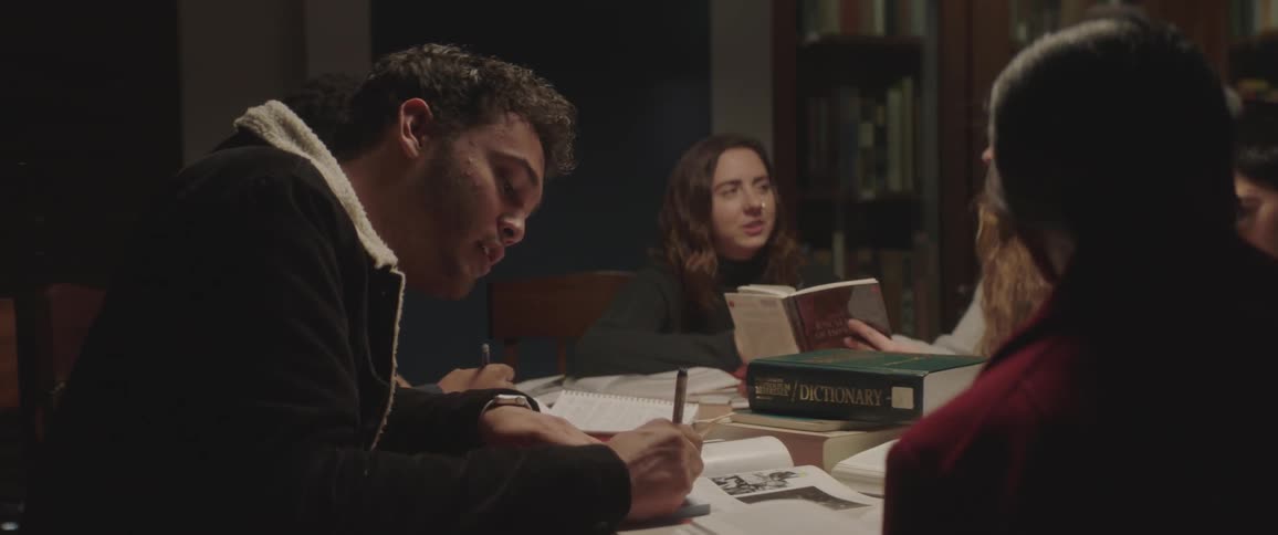 A group f male and female students are gathered around a table in a library with piles of books spread out before them