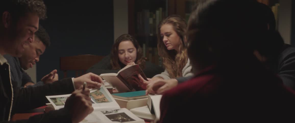 A group of teenagers sit at a table full of books talking while studying