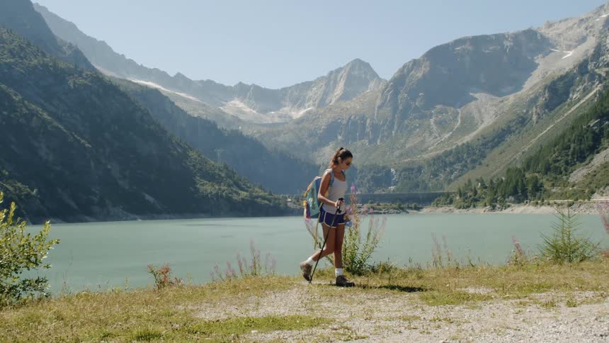 Woman holding hiking sticks walks next to the valley lake on a sunny day