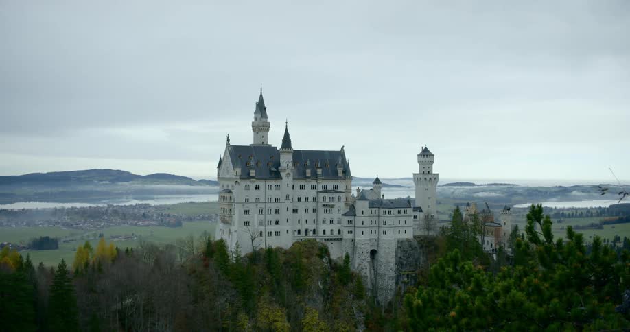 A beautiful castle with a tower and spires is located high above a dense forest and sprawling valley