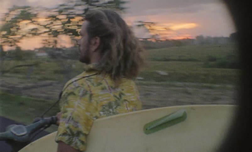 A male with long hair riding a moped while carrying a surfboard