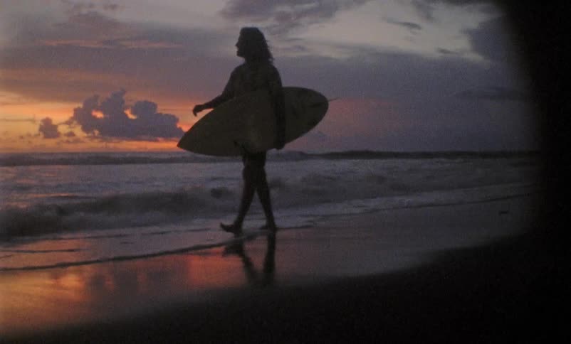 A man walks down a beach during sunset with a surfboard under his arm