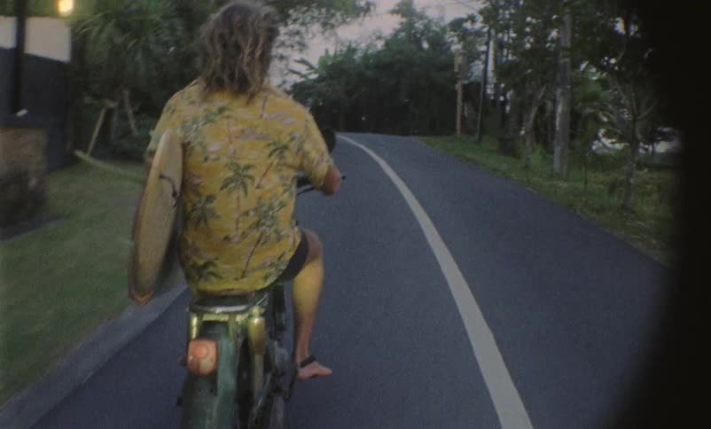 Man rides on a motorcycle down the street as he holds a surfboard under his arm