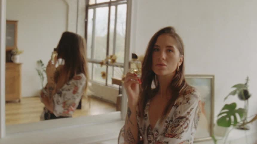 Woman looks in the mirror and shows perfume bottle indoors during daylight