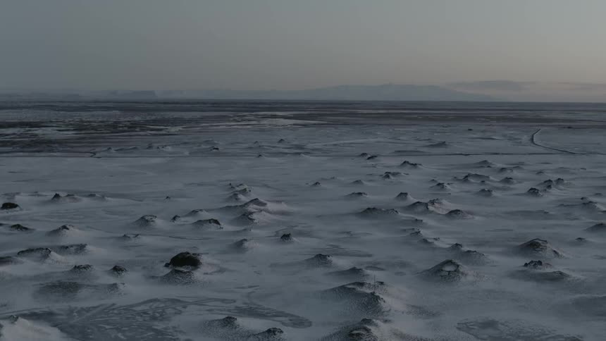 Snow covers frozen ice with mountains on the horizon under a cloudy sky