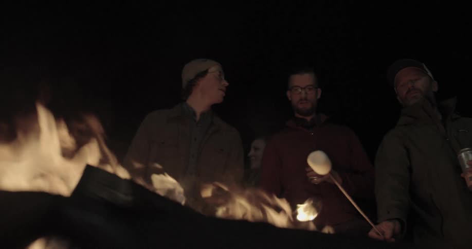 Men stand outside at night roasting marshmallows over bonfire