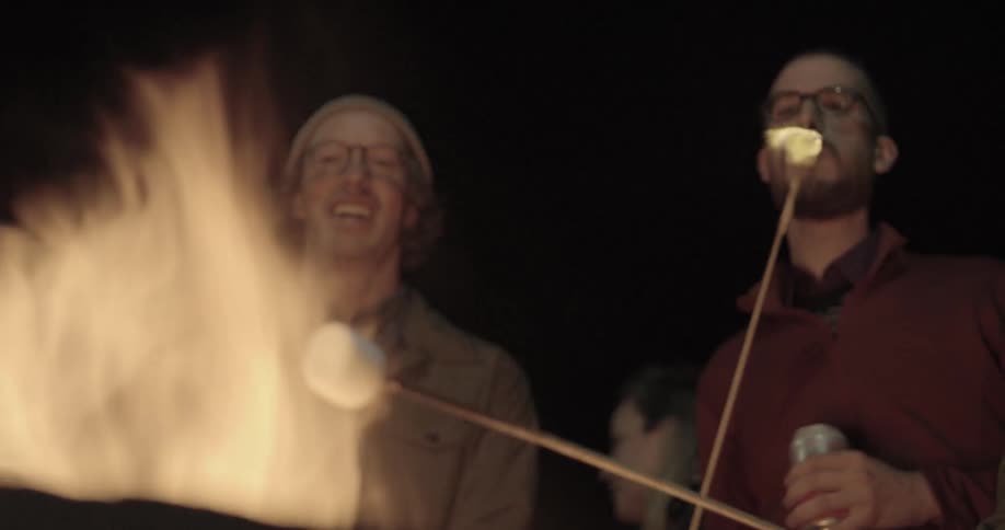 Two guys standing in front of a flame roasting marshmallows at night