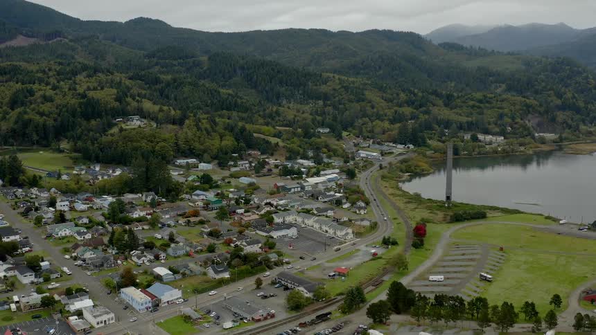 A small town with a number of cars driving through it sits at the base of a forest covered mountain located next to a lake