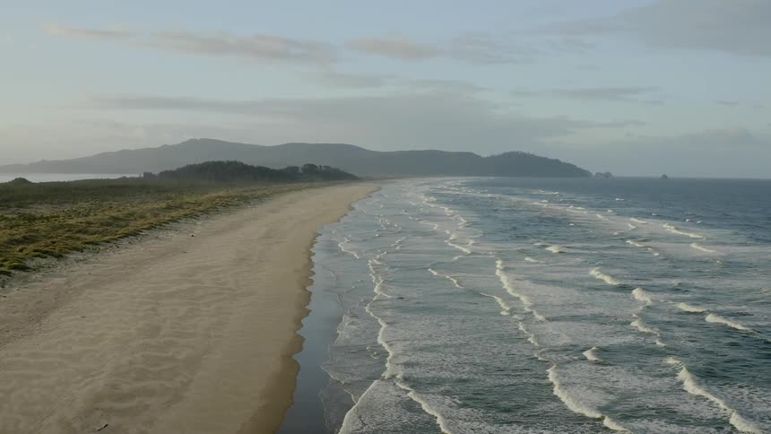 Row upon row of sets of waves can are washing towards a sandy beach shoreline as large mountains looms in the distance