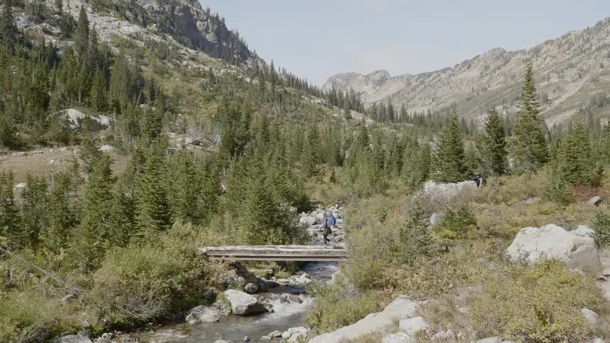 Man wearing a backpack walks over a bridge that is over a creek near the mountains