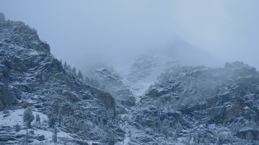 A treacherously rocky mountainous area is shrouded in a layer of snow and low hanging clouds