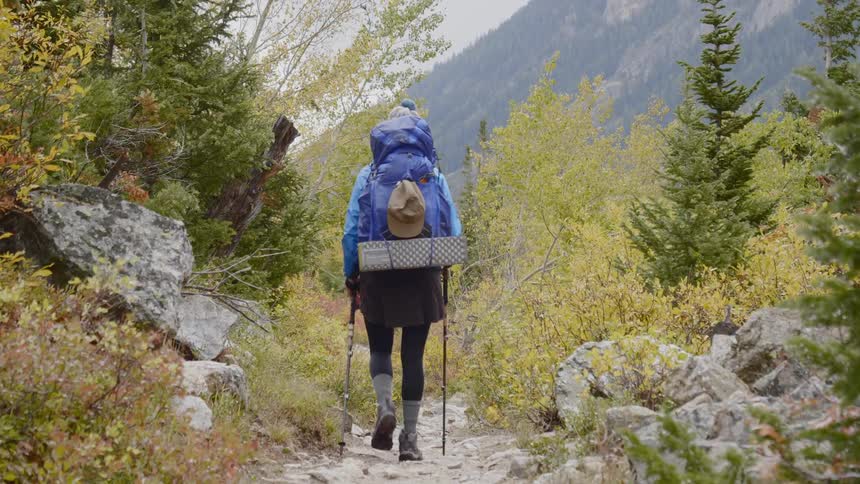 A female wearing an athletic skirt and large backpack is using hiking poles to navigate a narrow trail through the mountainous wilderness
