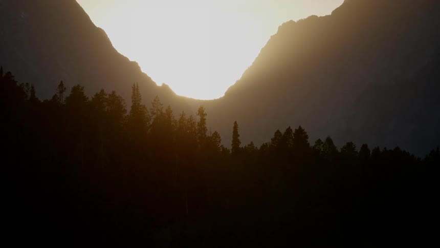 The sun shines from behind two mountains in a mountain range as the silhouette of a forest can be seen below