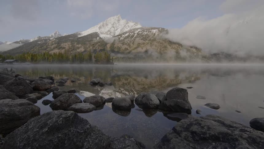 A tall snow capped mountain towers over a serene lake that has a thin fog hanging over the top of it