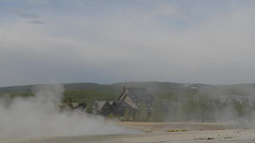 Steam billows up from the ground of a natural hot spring that is located in a clearing near a group of buildings located next to a forest