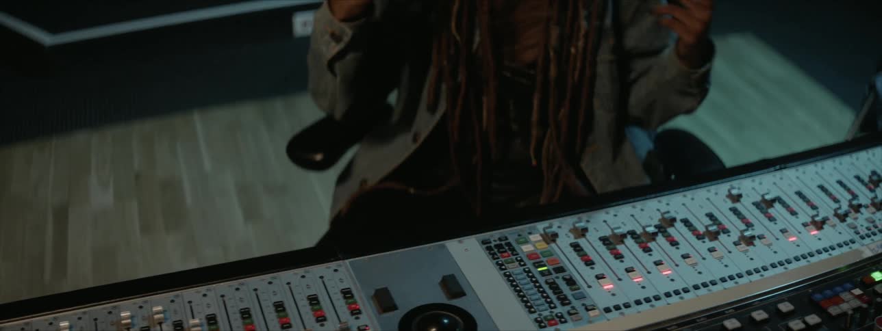 A black female with dreadlocks is dancing in her chair in front of a music soundboard which she makes a few adjustments to