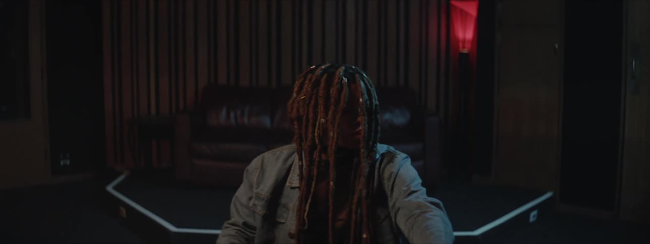 A black woman with long dreadlocks hanging in front of her face sits in front of a sound mixing board and bobs her head to music