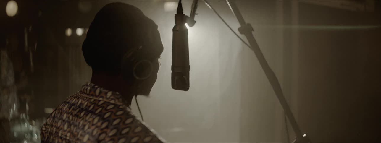 A man in a dark studio singing into a hanging microphone