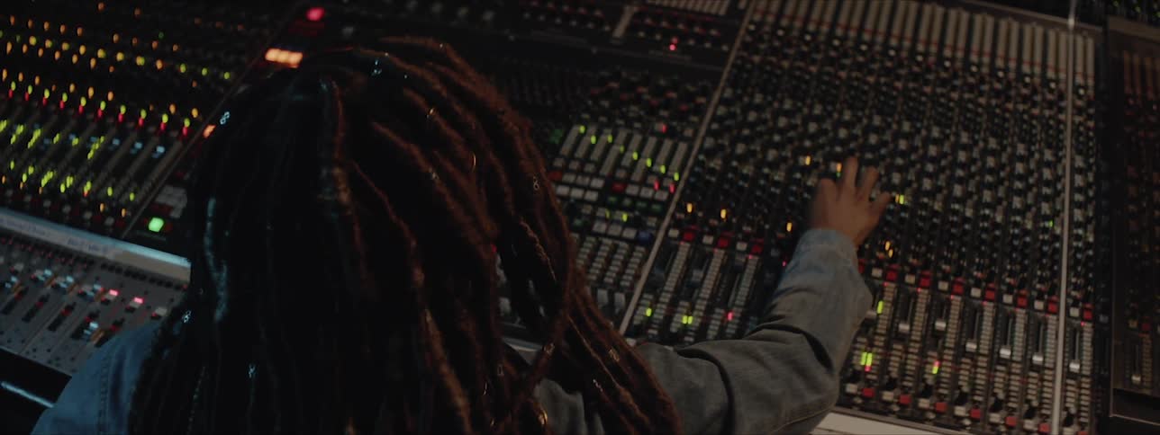 A black woman with long braids adjusts knobs on an enormous audio mixing board as she nods her head to music