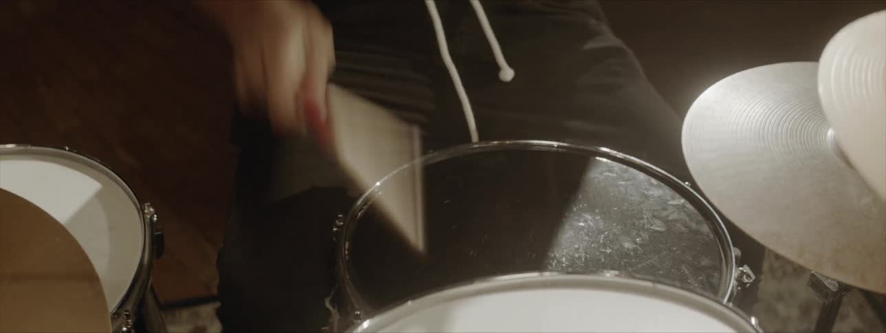 A musician sits at a drum set that he is playing by quickly striking the drums and cymbals with his drumsticks