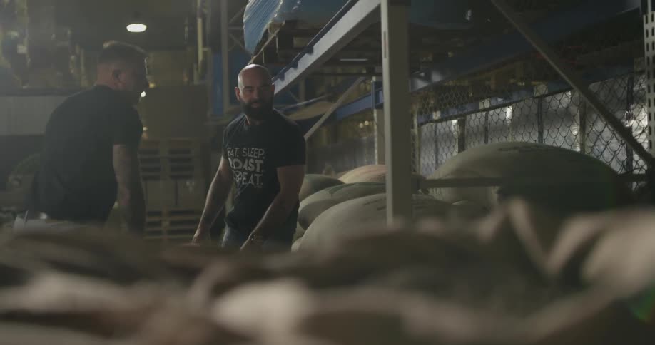 Two muscular men walk up a stack of huge burlap bags and together grab it by the corners and hoist it atop another stack of bags