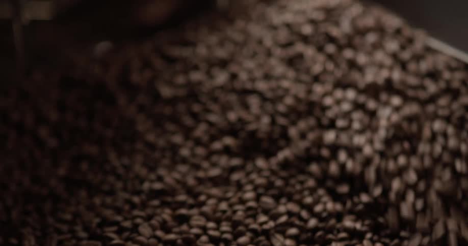 A huge container of whole coffee beans is being mixed and stirred by several metal mechanical arms that are sweeping by in a circular motion