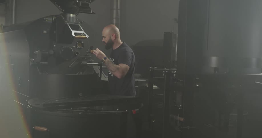 A bald man checks on the status of a large machine which he is using to roast coffee beans