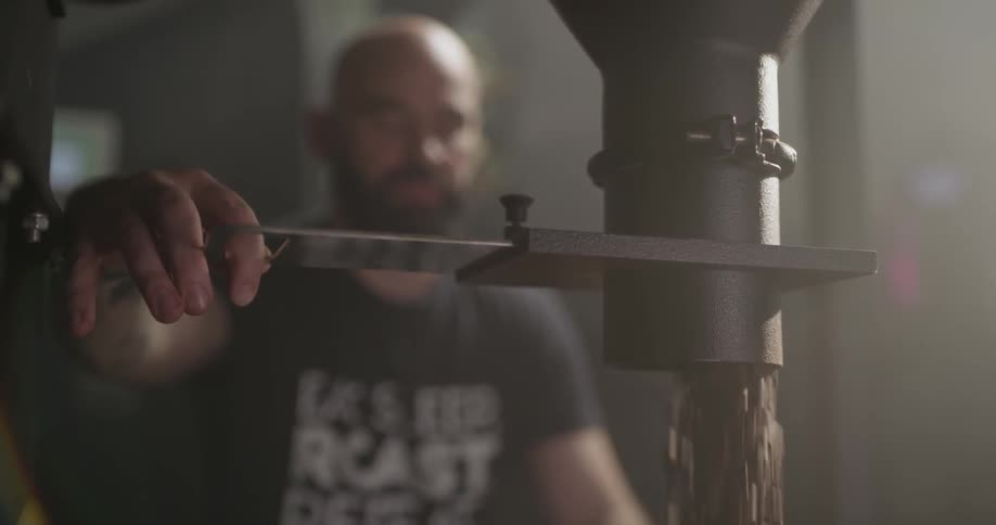 A bald man turns a handle on a machine and pours out a substantial number of coffee beans
