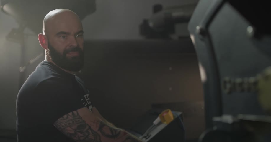 A bearded man sits in a room looking at machinery