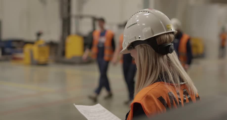 A woman in a hard hat and an orange vest reading a paper then waving to three men