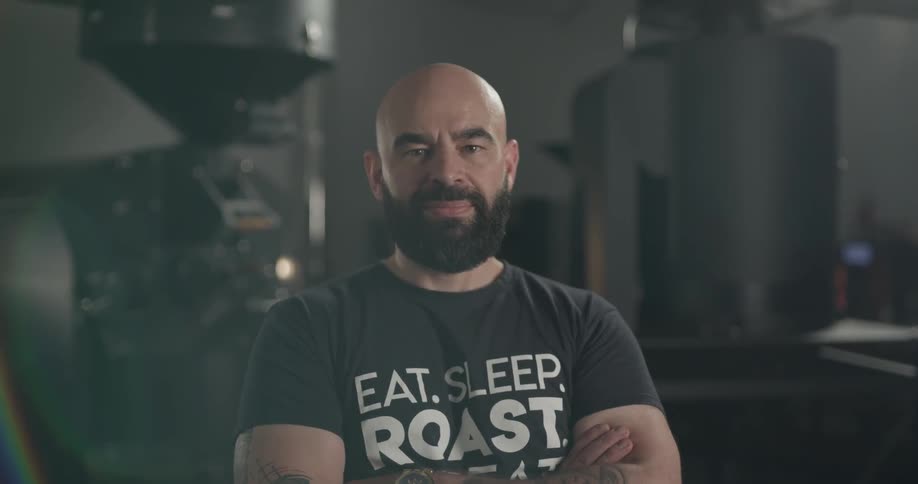 Bald bearded man stands cross armed looks at the camera indoors