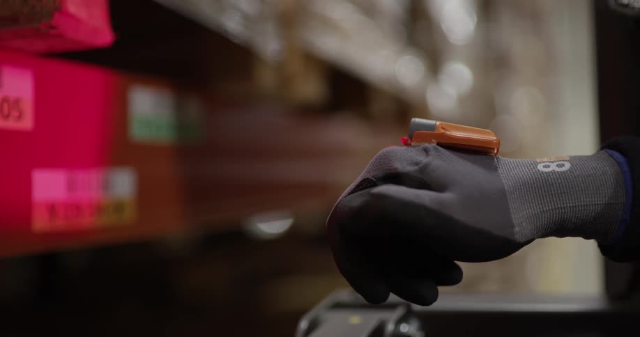 A person is wearing a glove with a light emitting device on the back of it that they are turning on and off while pointing it at some boxes on a shelf