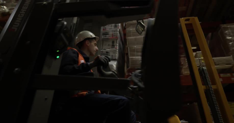 A man drives a forklift past tall metal shelving with boxes in a warehouse