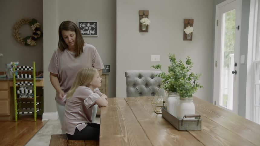 A mother brushes her daughters blonde hair at a dining table at home