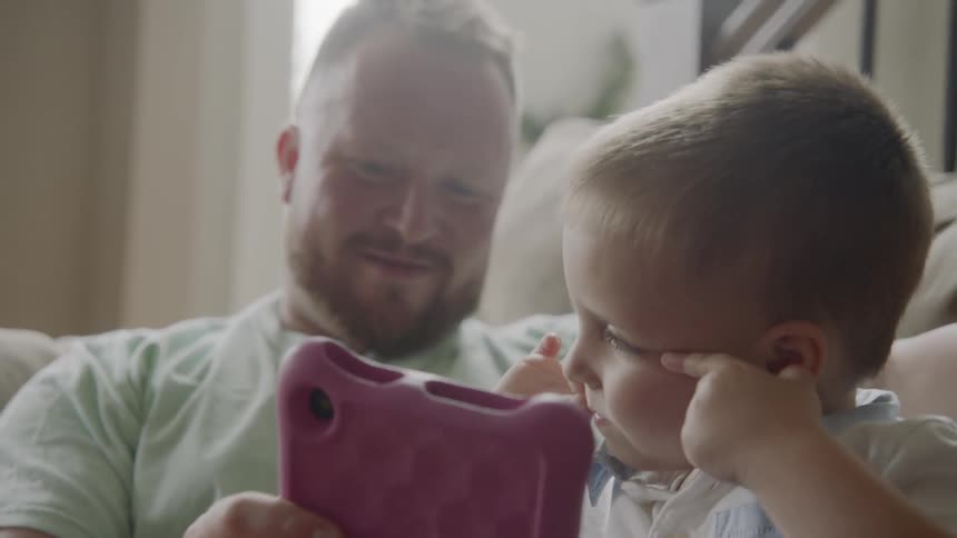Man sitting on the sofa uses tablet with toddler boy on a sunny day